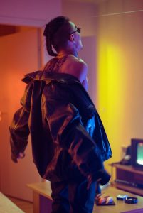 An edgy adult man with sunglasses and tattoos wearing a black leather jacket indoors.