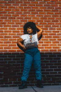 Stylish woman in fringed top and blue pants posing by a sunlit brick wall.