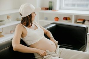 Pregnant woman in a white cap rests on a black sofa, showcasing her baby bump in a bright indoor setting.