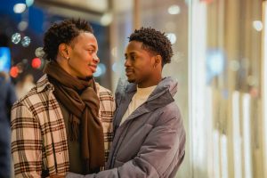 Romantic African American homosexual couple looking at each other while standing on street near building at evening time on blurred background