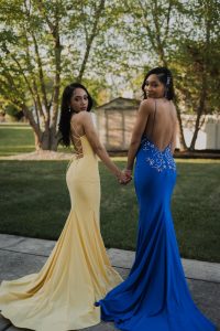Two women in elegant evening gowns pose outdoors during golden hour.