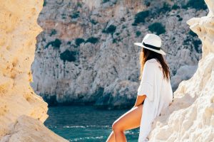 A woman in a hat sits on a rocky beach in summer, enjoying the sea view.