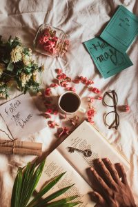 Top view of crop anonymous person with opened book and key placed on white textile with flowers and cup of drink near postcards with inscriptions