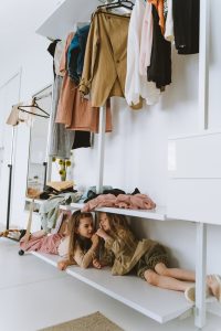 Two young girls playfully hiding under a closet shelf with clothes above.